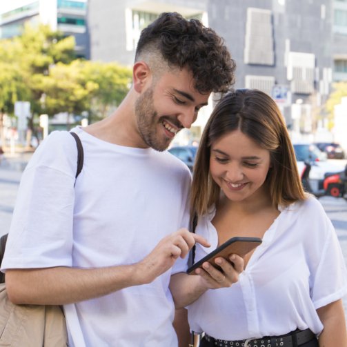Happy couple watching video on cell together in city square. Cheerful guy and girl standing outside, holding phone, looking at screen, laughing. Phone using concept