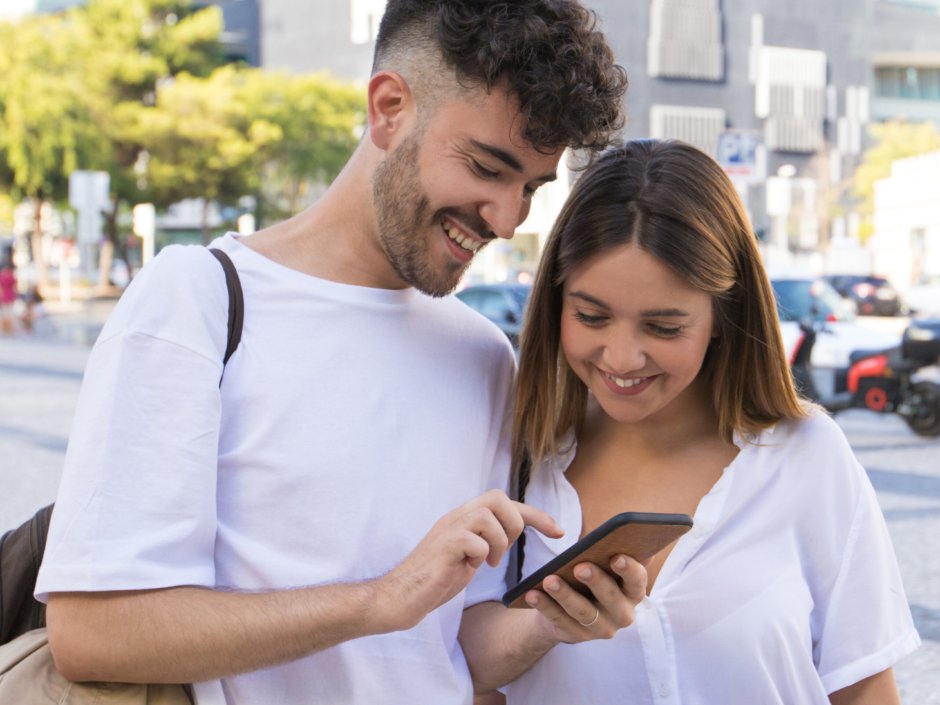 Happy couple watching video on cell together in city square. Cheerful guy and girl standing outside, holding phone, looking at screen, laughing. Phone using concept